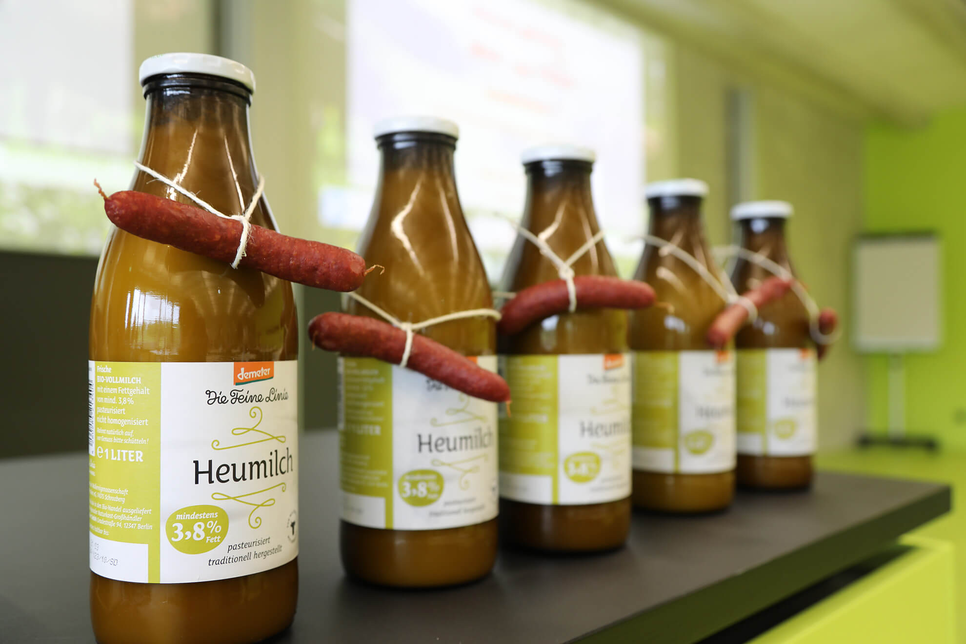 Flasche Demeter-Heumilch mit einer kleinen Wurst drumherum, um zu zeigen, dass mit jedem Liter Milch auch Rindfleisch entsteht.