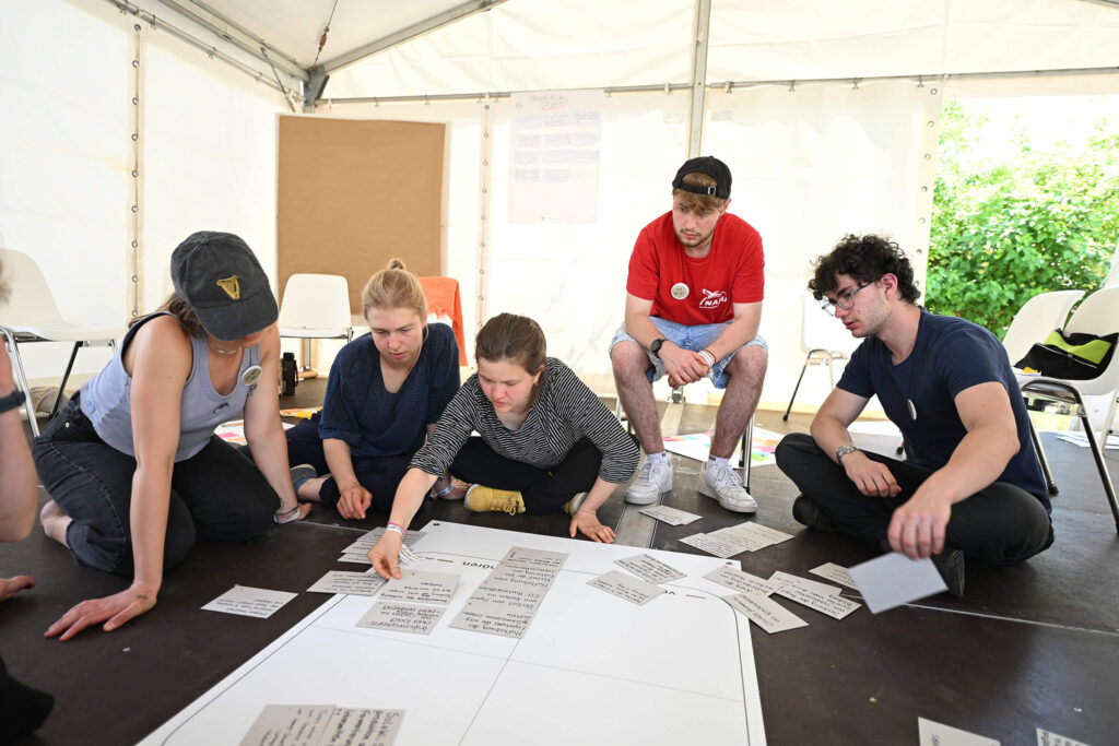Junge Männer und Frauen sitzen auf dem Boden und beschriften Karteikarten, die sie auf ein großes weißes Papier legen