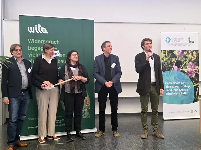 Eine Gruppe von vier Männern und einer Frau stehen vor einem Poster mit dem Auftruck "Wita" und halten einen Spaten.