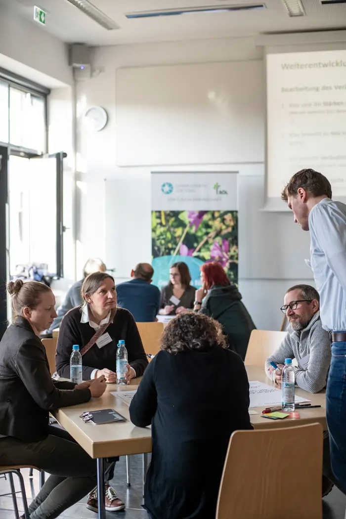 In einem Seminarraum sitzen Menschen um einen Tisch und sprechen miteinander, auf dem Tisch liegen Unterlagen zum Schreiben.
