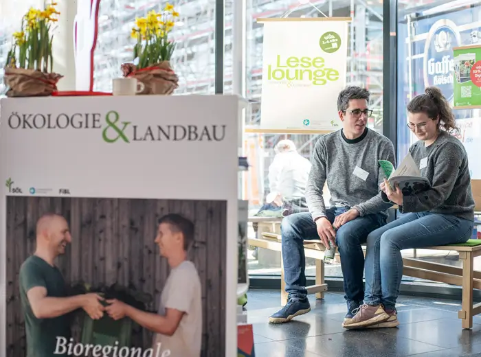 Ein junger Mann und eine junge Frau Ende 20 sitzen auf einer Holzbank vor einer Glaswand und lesen zusammen im Magazin Ökologie & Landbau. Beide Personen tragen Jeans, Turnschuhe, graue Pullover und eine Brille. Auf einem Plakat hinter den beiden steht das Wort Leselounge.